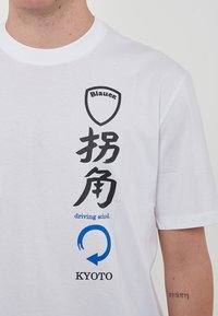 Maglietta in cotone bianco con un logo a scudo nero, caratteri giapponesi e la scritta "scuola di guida" in blu, con "KYOTO" sottostante.