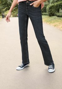 Ciemnoniebieskie dżinsy w stylu dzwonów, wysokie w talii z przednimi kieszeniami. Noszone z granatowymi trampkami z białymi akcentami i sznurówkami, ustawione na ścieżce.