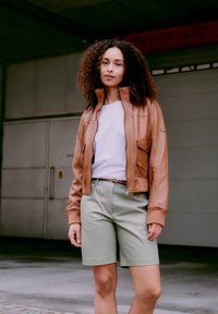 Kvinne med krøllete hår iført en brun skinnjakke, hvit skjorte og lysegrønne knelange shorts som står foran en lukket garasjeport.