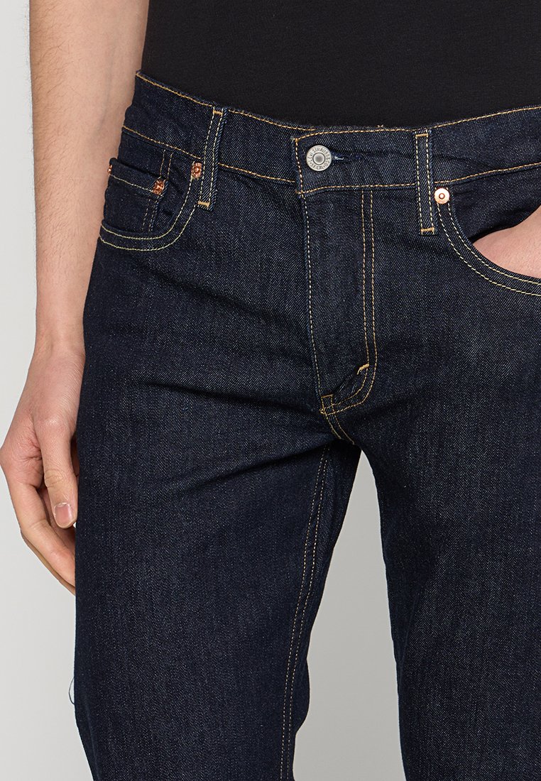 Jean en denim bleu foncé avec des coutures jaunes, design à cinq poches, boutons en métal et rivets, et coupe slim.