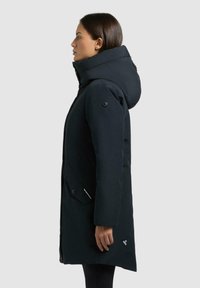Schwarze Daunenjacke mit einer großen Kapuze, Knopf- und Reißverschluss, seitlichen Taschen und Logodetail am Saum. Schlichte, matte Oberfläche.