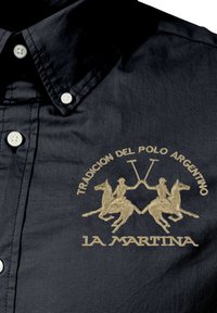 Czarna koszula z tkaniny z kołnierzem na guziki. Złote hafty przedstawiają dwa konie z jeźdźcami oraz napisy "Tradicion del Polo Argentino" i "La Martina."