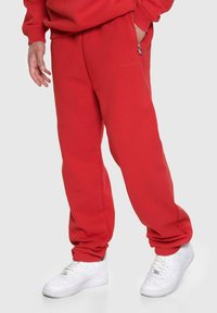 Rote Sweatpants aus weichem Material, mit einer Reißverschlusstasche und gerippten Bündchen. Kombiniert mit weißen Sneakern. Einfaches Logodetail am Bein.