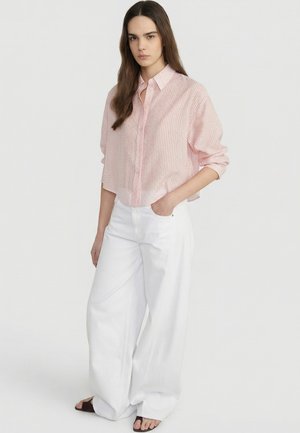 Kobieta o długich ciemnych włosach, ubrana w jasnoróżową koszulę w paski zapinaną na guziki oraz szerokie białe spodnie, stoi z jedną ręką w kieszeni.