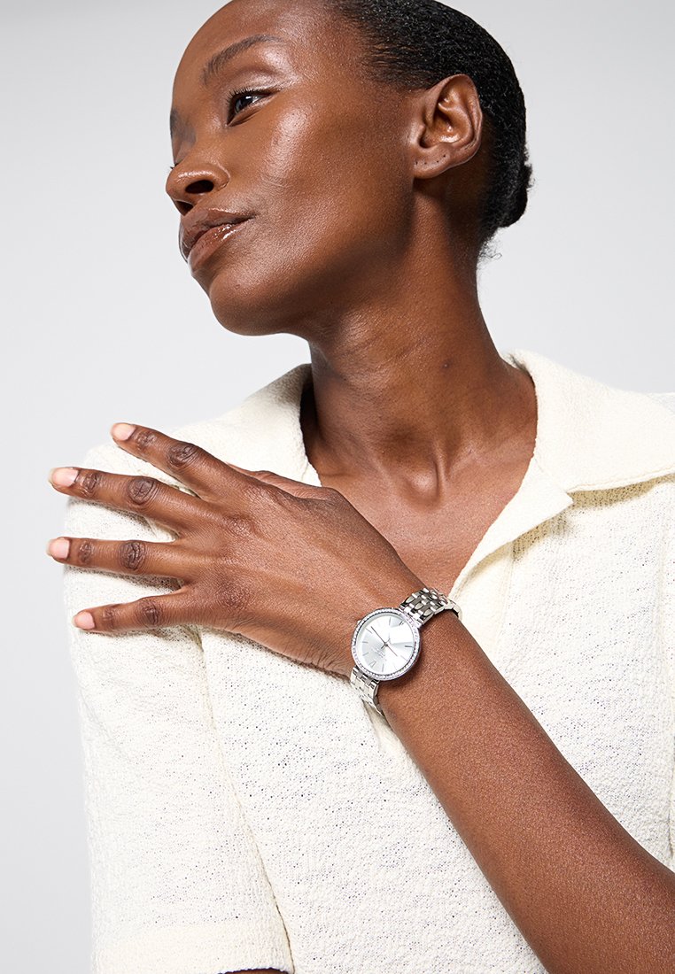 Montre-bracelet en argent avec un cadran rond blanc, entouré d'une lunette texturée, et un bracelet à maillons métalliques. Stylée avec un haut texturé de couleur claire.