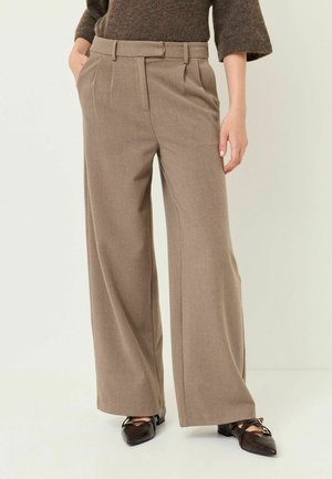 Person trägt hoch taillierte, weite beige Hose mit Falten, braunen Pullover und dunkelbraune spitze Loafers, steht mit den Händen in den Taschen.