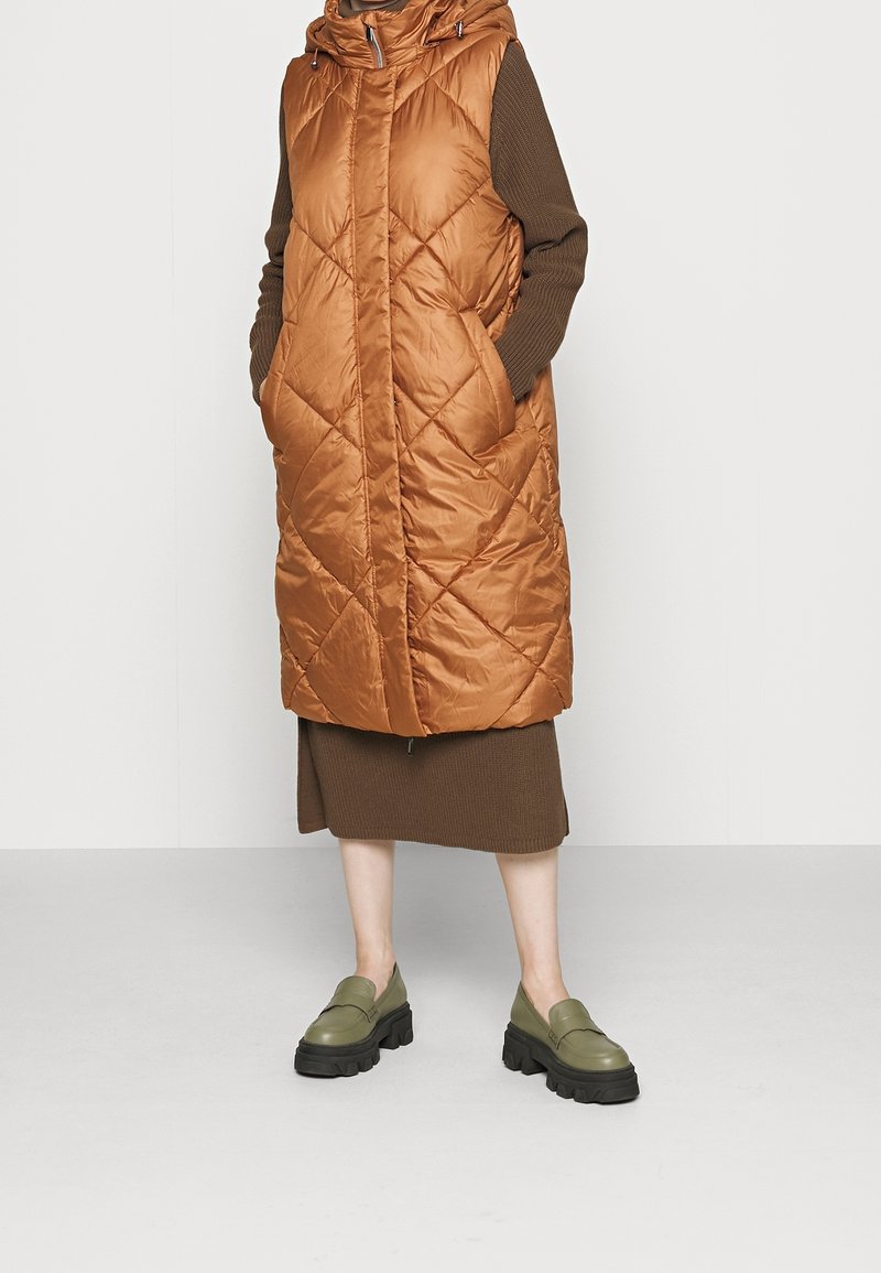 Gepolsterte, ärmellose Jacke in glänzendem Rostorange mit Rautenmuster, getragen über einem braunen, gerippten Kleid. Die Schuhe sind klobige, olivgrüne Loafers.