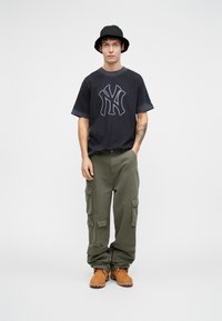 Camiseta preta de manga curta com um logo cinza "NY", calças cargo verdes com vários bolsos e botas em tom areia. O modelo usa um chapéu à beira preta.