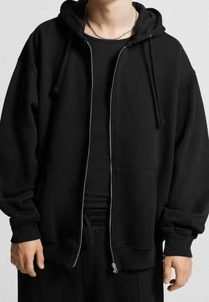 Person trägt einen teilweise geschlossenen schwarzen Hoodie mit Zugbändern über einem schwarzen gerippten Shirt und schwarzen Hosen vor einem grauen Hintergrund.