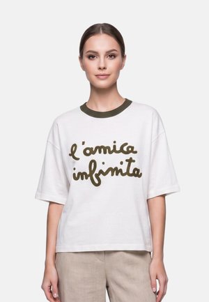 Femme aux cheveux bruns en chignon bas porte un t-shirt blanc avec un col vert foncé et le texte "l'amica infinita", associé à un pantalon beige.