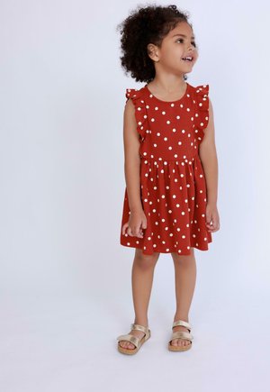 Czerwona sukienka w białe kropki z falbaniastymi rękawami, dopasowanym topem i rozkloszowanym dołem. Zestawiona ze złotymi sandałami na gołych stopach. Prosty i zabawny wzór.