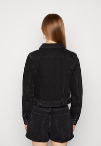 Kvinna med långt rakt hår klädd i svart denimjacka och matchande höga shorts, stående med ryggen mot kameran.