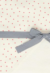 Coperta in cotone con sfondo crema, decorata con piccoli cuori rossi e un fiocco a righe blu e bianche in un angolo.