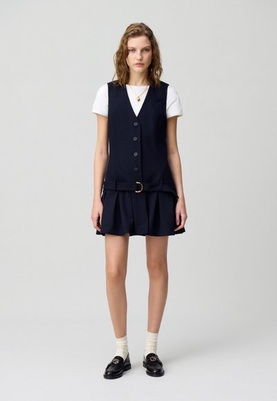 Robe bleu marine avec un corsage cintré, des boutons sur le devant et une jupe plissée. Portée sur un t-shirt blanc, associée à des mocassins noirs et des chaussettes crème.
