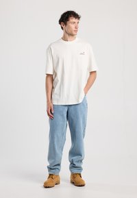 Kerman puuvillapaita, jossa on pyöreä kaula-aukko, lyhyet hihat ja pieni logo rinnassa, yhdistettynä vaaleansinisiin väljiksi ommeltuihin farkkuihin ja keltaisiin saappaisiin.