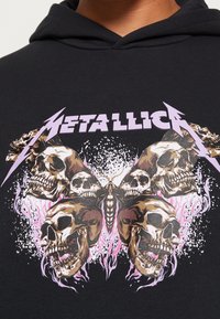 Černá mikina s kapucí s grafickým designem stylizovaných lebek a motýla v odstínech hnědé a růžové, s nápisem "METALLICA" v purpurovém textu nahoře.