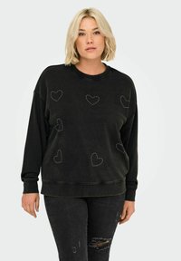 Svart sweatshirt med avslappnad passform, rund halsringning och sydda hjärtdesigner i silver nitar över framsidan. Slitna jeans kompletterar outfiten.