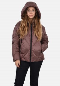 Giacca imbottita marrone trapuntata con cappuccio, dotata di chiusura frontale con cerniera e tasche laterali. Il modello è ritratto con le mani nelle tasche, indossando jeans scuri.