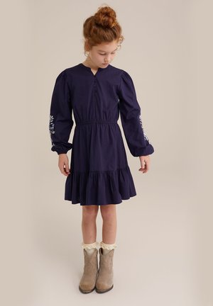 Robe longue à manches longues bleu marine avec patte de boutonnage et ourlet à volants, ornée de broderies florales sur les manches, portée avec des bottines beige et des chaussettes en dentelle blanche.