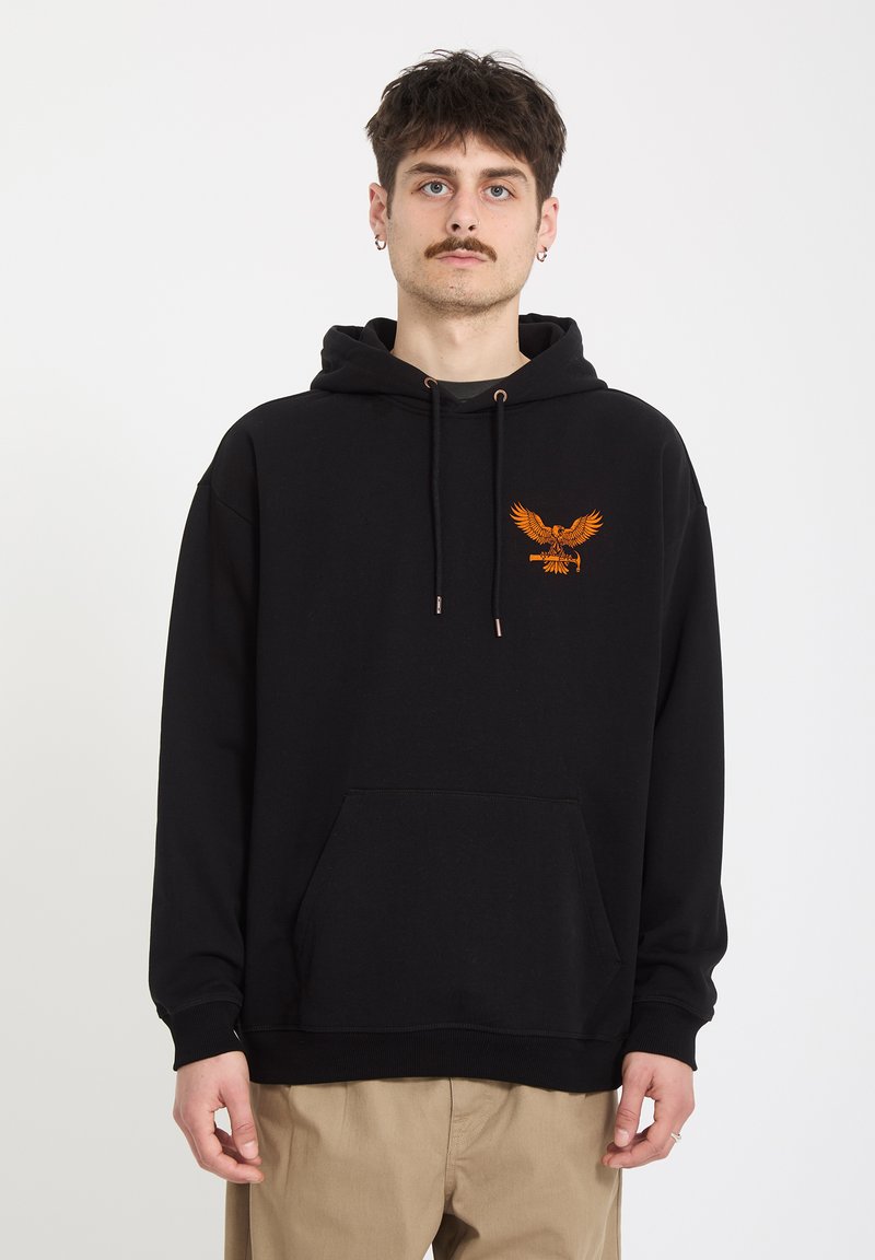 Jeune homme aux cheveux courts et foncés avec une moustache, portant un sweat à capuche noir orné d'un aigle orange sur la poitrine et un pantalon beige, debout devant un fond blanc.