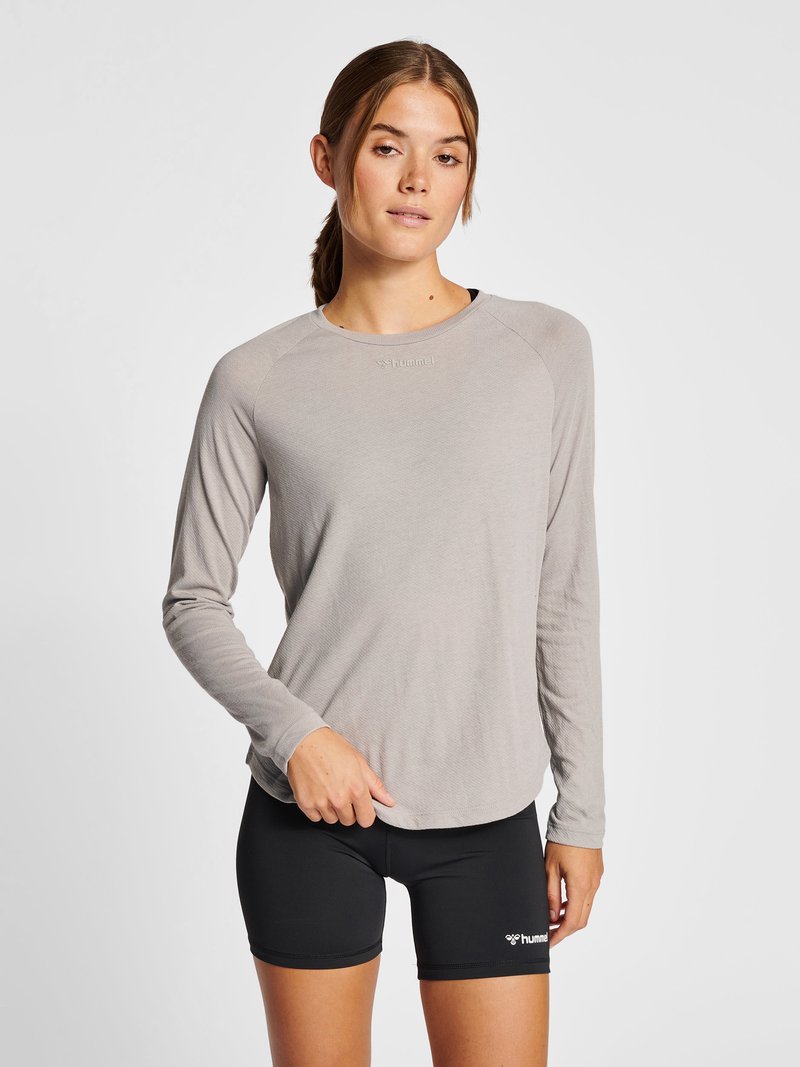 Vrouw draagt een lichtgrijze lange mouwen top en zwarte sportshorts, staand tegen een effen achtergrond.
