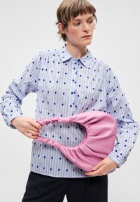 Ροζ συνθετική τσάντα ώμου με λεπτομέρειες πτυχώματος, κρατημένη σε χέρι, συνδυασμένη με μπλε και λευκό ριγέ πουκάμισο με υφή πουά.