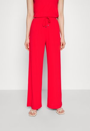 Femme portant un pantalon rouge taille haute, jambes larges, avec cordon de serrage et des sandales blanches à bouts ouverts, debout devant un fond uni.