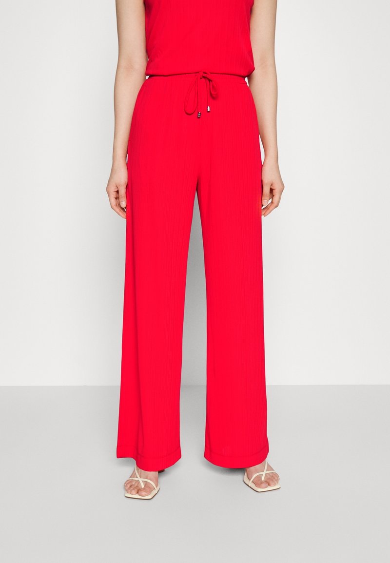 Femme portant un pantalon rouge taille haute, jambes larges, avec cordon de serrage et des sandales blanches à bouts ouverts, debout devant un fond uni.