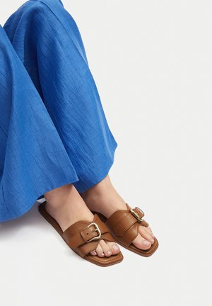 Brązowe skórzane klapki na płaskiej, prostokątnej podeszwie z szerokim paskiem i złotą klamrą. Podeszwa pokryta jest niebieskim, teksturowanym materiałem.