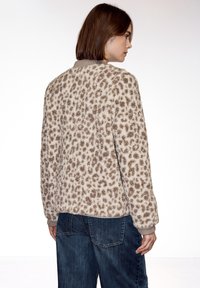 Femme aux cheveux bruns courts portant une veste pelucheuse à imprimé léopard beige et marron et un jean bleu, légèrement tournée de côté par rapport à la caméra.