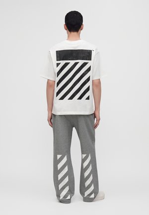 Mann trägt weißes T-Shirt mit schwarzem diagonalem Streifenblock und graue Hose mit weißen diagonalen Streifen, steht mit dem Rücken zur Kamera.