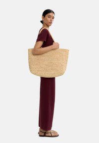 Duża pleciona torba ze słomy o miękkim, zaokrąglonym kształcie i grubymi plecionymi rączkami. Modelka ma na sobie burgundową dopasowaną sukienkę i sandały na cienkich paskach.