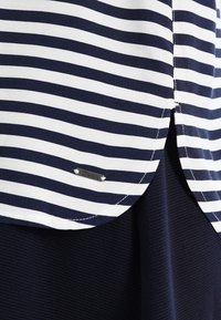 Tkanina w poziome paski w kolorze granatowym i białym, z zaokrąglonym dołem, na gładkiej granatowej, prążkowanej tkaninie, z małą metalową metką "Denim".