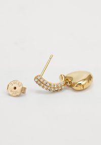 Okrugle naušnice zlatne boje s zakrivljenim dizajnom ukrašenim prozirnim rhinestones. Uključuju okruglu pozadinu s natpisom "Guess".
