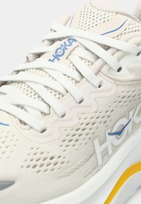 Scarpa atletica beige chiaro con tomaia in rete, lacci bianchi, dettagli blu e una suola bianca chunky con un particolare giallo.
