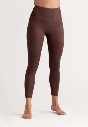 Frau trägt hoch taillierte, knöchellange dunkelbraune Leggings und steht barfuß vor einem einfachen hellen Hintergrund.