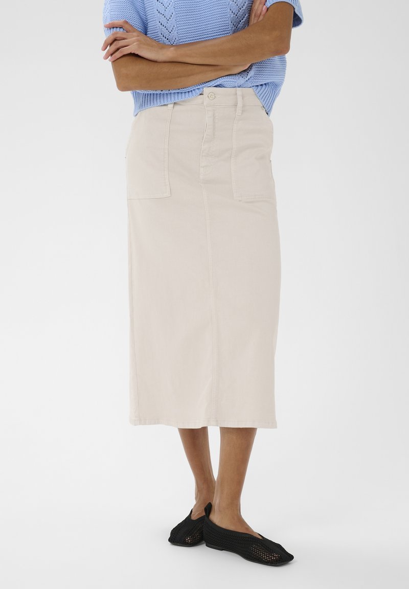 Personne portant une jupe beige mi-longue, un haut en maille bleu clair à manches courtes et des chaussures noires en filet sans lacets, debout les bras croisés.