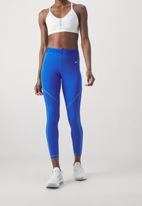 Nike Sport-BH in Weiß mit grauen Akzenten, kombiniert mit blauen Nike Leggings mit kontrastierenden Nähten. Leichter, elastischer Stoff, figurschmeichelndes Design.