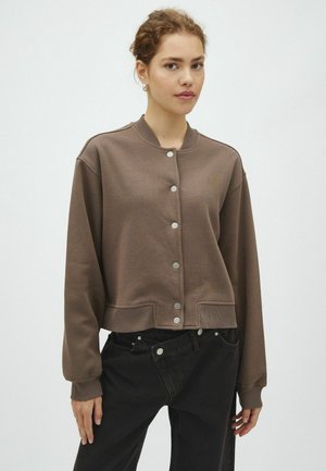 Jeune femme aux cheveux bouclés porte une veste marron boutonnée et un pantalon noir sur un fond clair uni.