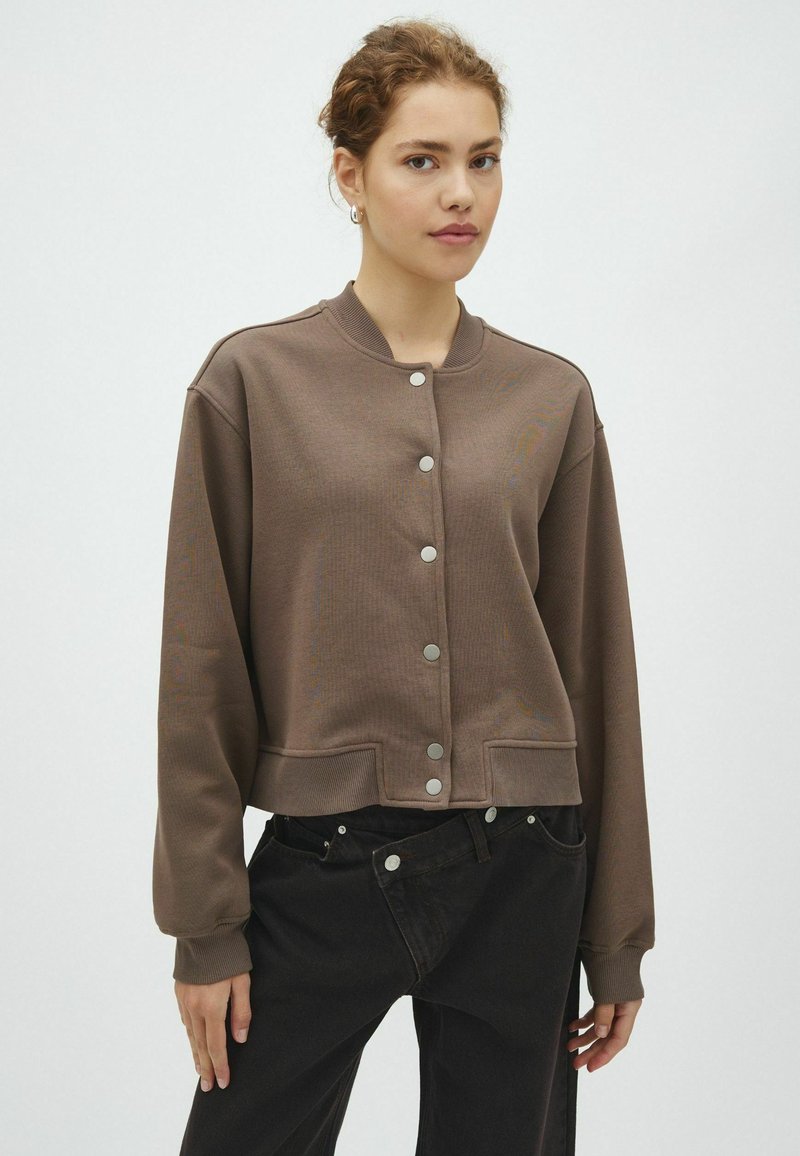 Jeune femme aux cheveux bouclés porte une veste marron boutonnée et un pantalon noir sur un fond clair uni.