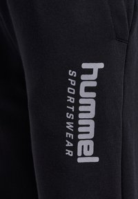 Zwarte sportbroek van zacht materiaal met een verticale witte "hummel" logo en "SPORTSWEAR" tekst langs de zijkant.