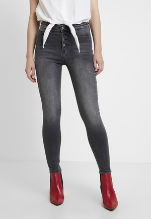 Osoba ubrana w wyblakłe czarne rurki z wysokim stanem i przednimi guzikami, białą związaną koszulę oraz czerwone botki stojąca na białej podłodze.