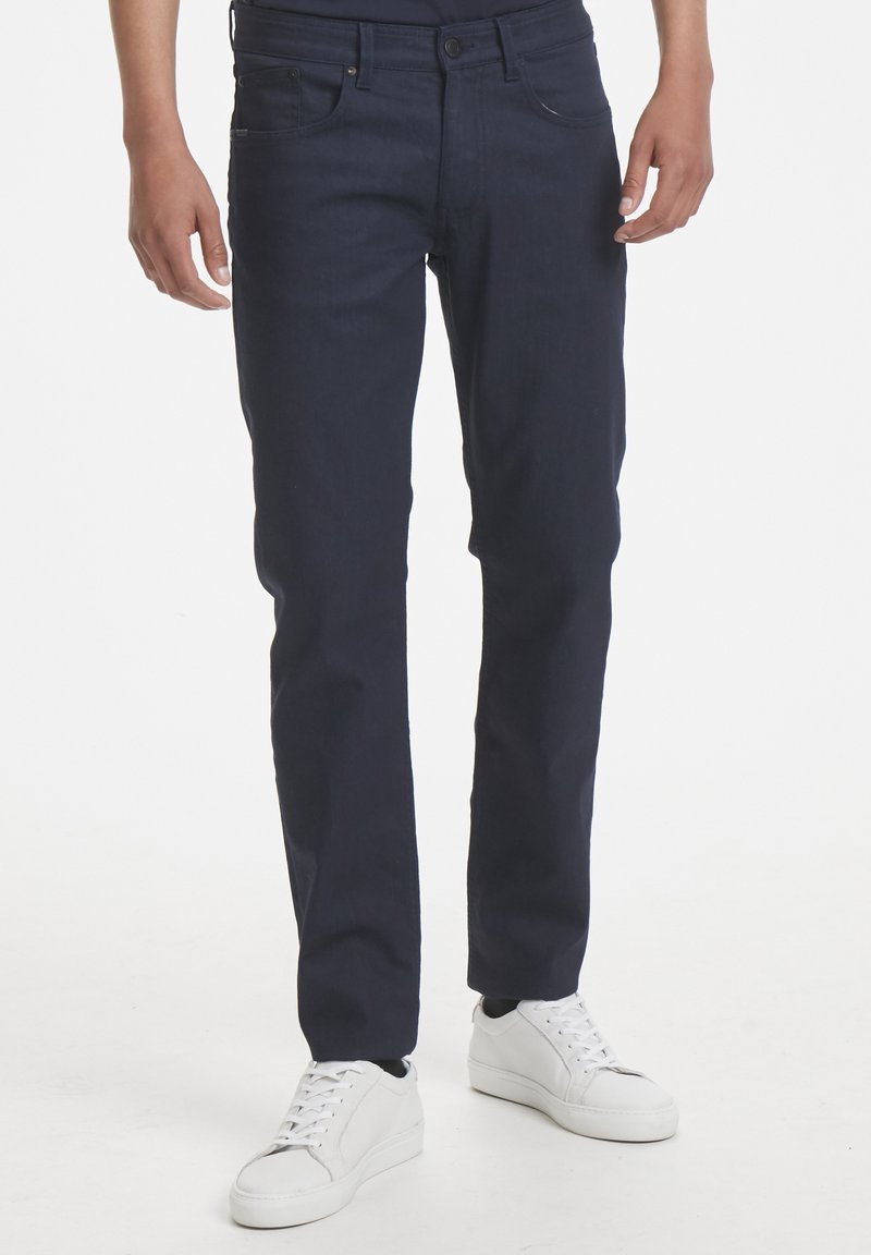 Jeans en denim bleu marine foncé à coupe slim, dotés d'un design standard à cinq poches et d'une fermeture à boutons. Portés avec des baskets blanches.