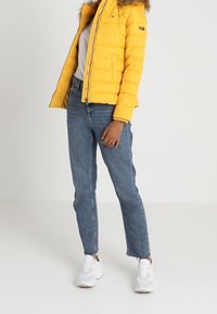 Żółta kurtka puchowa z kapturem z sztucznego futra, zestawiona z niebieskimi dżinsami i białymi sneakersami. Kurtka ma zamek błyskawiczny z przodu i kieszenie.