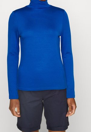 Persona que lleva una camiseta de cuello alto ajustada de manga larga en azul real y pantalones cortos negros hasta la rodilla sobre un fondo sencillo.