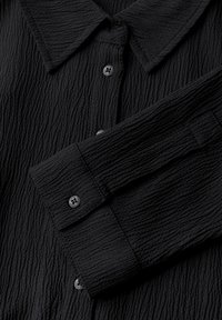 Camicia nera in tessuto texturizzato con colletto a punte, parte anteriore con bottoni e maniche con polsini dotati di bottoni circolari. Il tessuto presenta un motivo sgualcito.