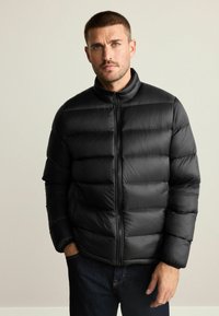 Sort pufferjakke med høj krave, horisontalt kviltet design, lynlås foran og en glat, blank overflade. Båret over mørke jeans.