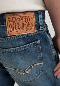Ljusblå denimjeans med en läderetikett som säger "Replay Blue Jeans." Har kontrasterande sömmar, bakfickor och en sliten textur.