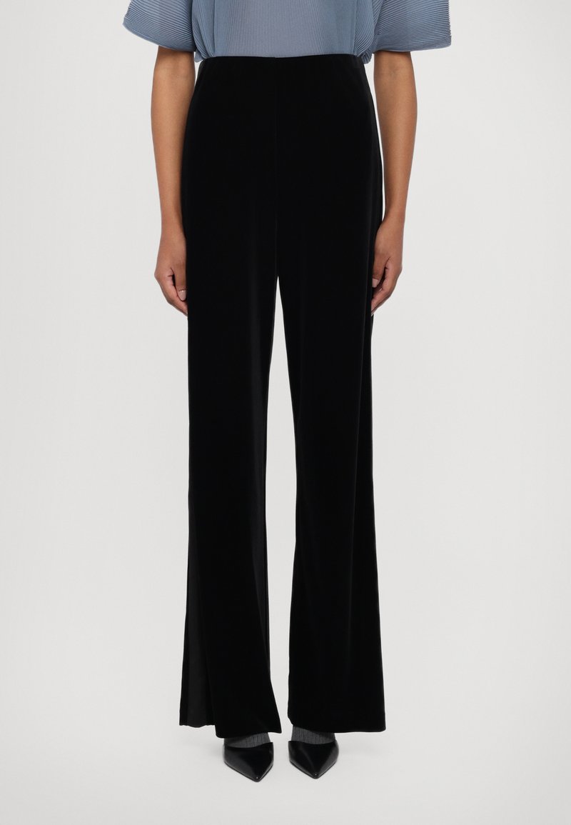 Pantalons larges en velours noir avec une texture lisse. Présentent une taille haute et sont associés à un haut plissé bleu clair et des talons aiguilles.