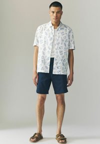 Chemise à manches courtes blanche avec des motifs de fruits et de boissons bleus, associée à un short bleu marine et des sandales marron. Design simple et coupe décontractée.